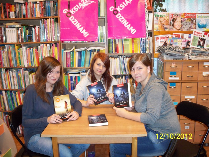 3 dziewczyny siedzące przy kwadratowym stoliku, w rękach trzymają przeczytane książki i pokazują ich okładki w stronę aparatu