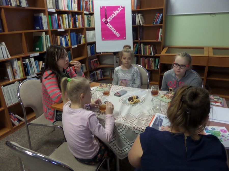 5 dziewczynek siedzi przy stole, jedzą ciastka, piją herbatę, rozmawiają oprzeczytanej książce.