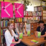 Trzy osoby, dwie nastolatki i jedna kobieta siedzą przy stole, patrzą w obiektyw aparatu, pokazują okładki książek trzymanych w rękach.