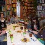 Osiem dziewczyn siedzących wokół stołu, część czyta książki, inne słuchają lub rozmawiają