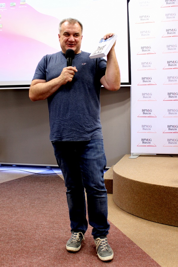 Marcin Kostrzyński, postawny mężczyzna o siwych włosach ubrany w granatową koszulkę, spodnie jeansowe i sportowe buty, stoi uśmiechnięty, w jednej ręce trzyma mikrofon w drugiej książkę.