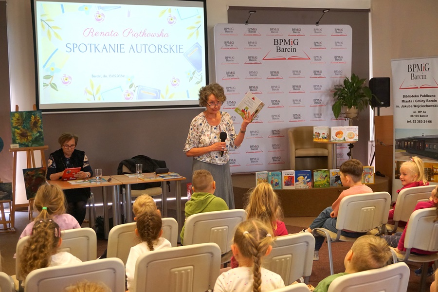 Zaproszona autorka trzymając książkę w ręku patrzy w stronę dzieci