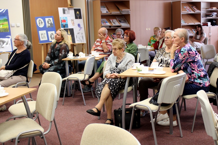 kobiety i meżczyźni siedzący przy stolikach słuchający rozmowy bibliotekarki prowadzącej spotkanie i zaproszonego autora