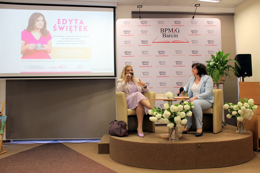 Z prawej strony na scenie siedzi Renata Grabowska i Edyta Świętek, kobiety rozmawiają ze sobą o twórczosci autorki, po lewej stronie zdjęcia widać wyświetlany na ekranie projektora plakat promujący temat spotkania.