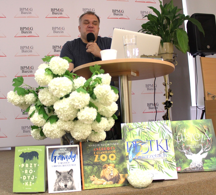 Pan Marcin siedzi przy laptopie, przed nim na scenie ustawione są książki jego autorstwa, oraz kwiaty w wazonie