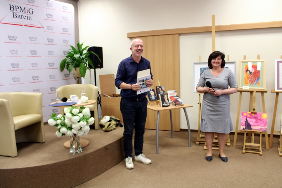 po prawej stronie dyrektorka biblioteki w Barcinie, po lewej zaproszony gość Michał Wójcik, obydwoje sie uśmiechają, pan Michał trzyma w rękach wręczone podziękowania za spotkanie i upominek w formie książki