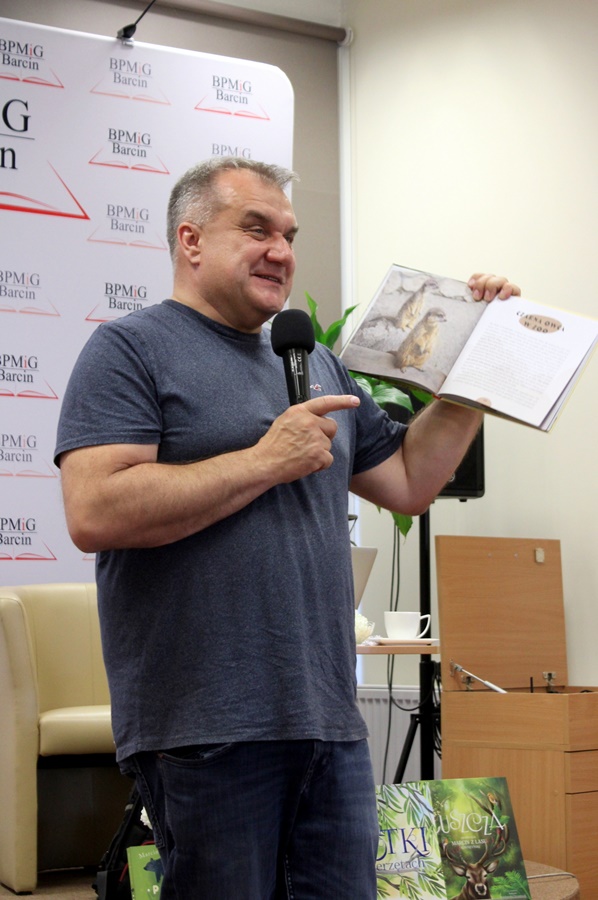 Pan Marcin Kostrzyński stoi przed publicznością i wskazuje na książkę trzymaną w jednej ręce.