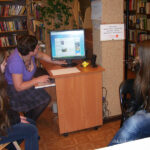 bibliotekarka siedzi przy komputerze, monitor odwrócony jest w stronę 2 dziwczyn obecnych na spotkaniu na monitorze wyświetlana jest jakaś strona www