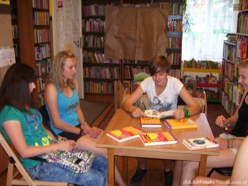 4 osoby przy kwadratowym stole, jedna pochyla się nad otwartą książką