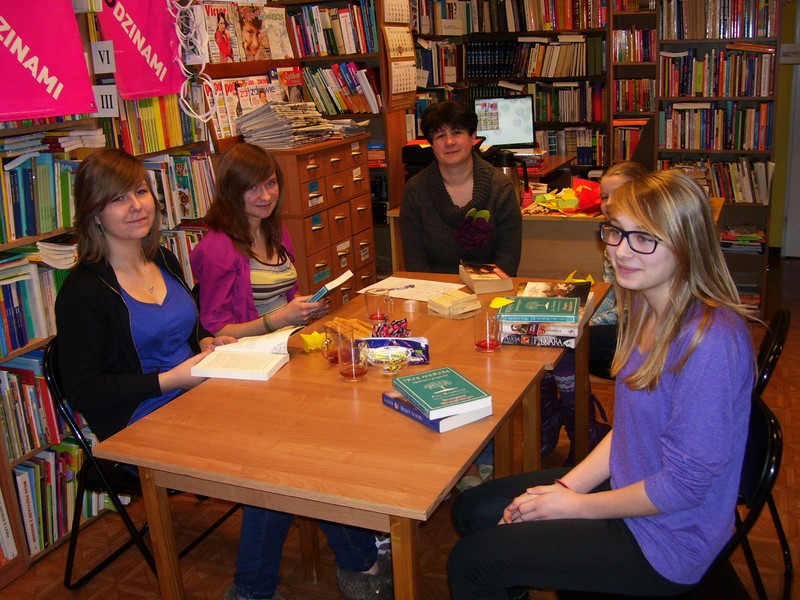 5 osó bsiedzących przy stole na którym leżą książki, talerzyki z ciaskami, stoją szklanki