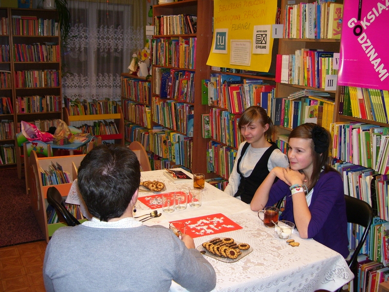 Trzy osoby siedzą przy stole i rozmawiają między sobą, na stole leżą książki, stoją szklanki i talerzyki z ciastkami