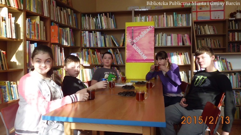Grupa dzieci siedząca przy stołach, pije herbatę, na talerzykach leżą ciastka, w tle widać plakat z napisem godzinami o książkach.