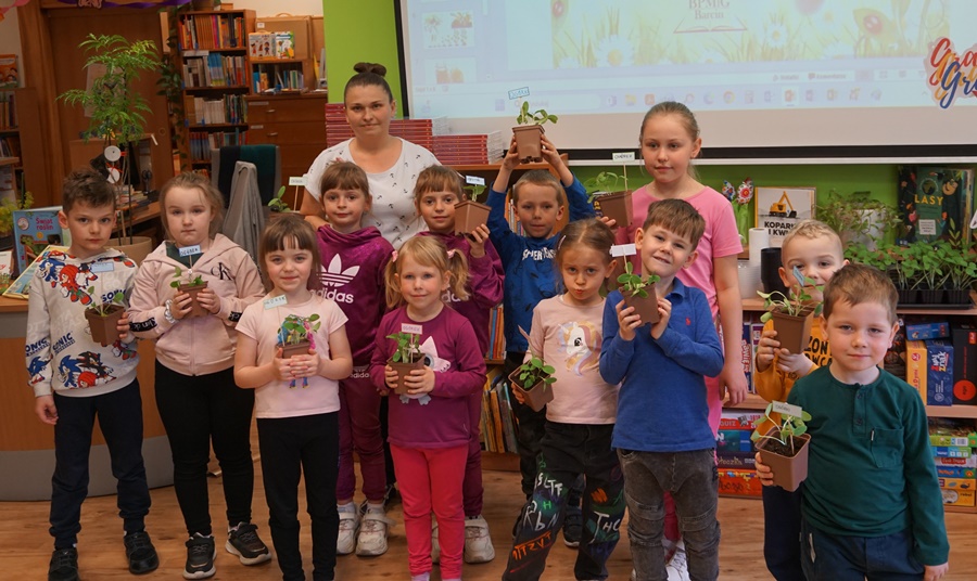grupowe zdjęcie dzieci biorących udział w warsztatach, razem z dziećmi stoi bibliotekarka, dzieci trzymają doniczki z sadzonkami