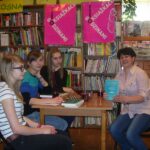 uczestniczki spotkania i bibliotekarka siedzą przy stole, 3 osoby patrzą w stronę aparatu jedna patrzy przed siebie, prowadząca spotkani pokazuje okładkę książki