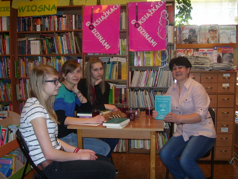 uczestniczki spotkania i bibliotekarka siedzą przy stole, 3 osoby patrzą w stronę aparatu jedna patrzy przed siebie, prowadząca spotkani pokazuje okładkę książki