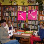 3 osoby siedzące przy stoliku trzymające w rękach książki, na stole leżą kolorowe kartki papieru na których widać białe papierowe paski
