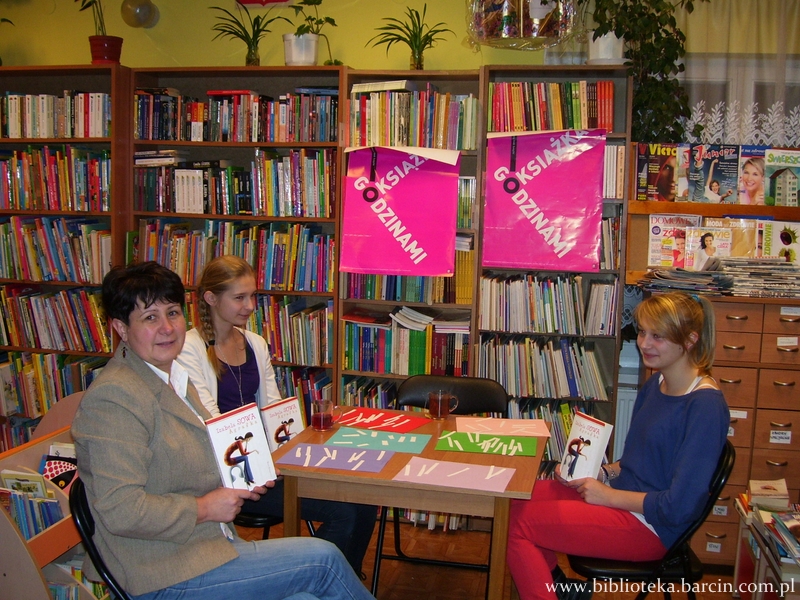 3 osoby siedzące przy stoliku trzymające w rękach książki, na stole leżą kolorowe kartki papieru na których widać białe papierowe paski