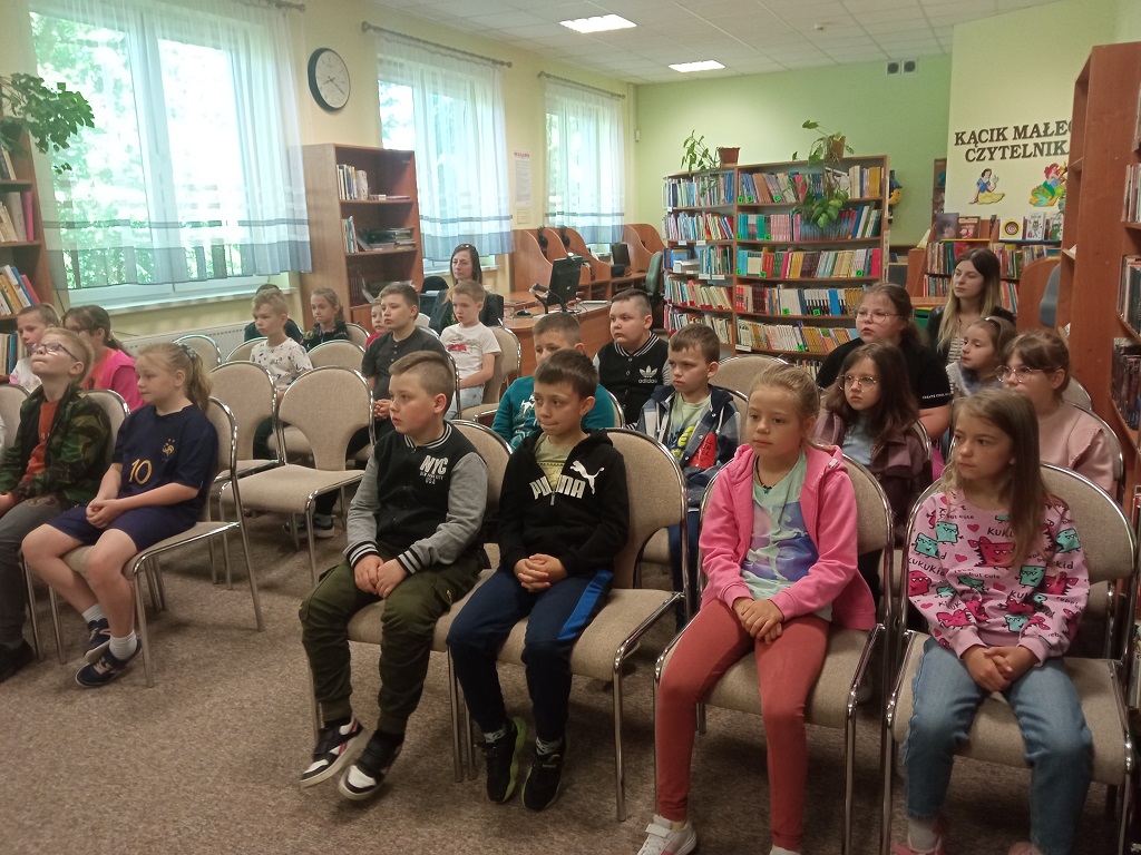 Zebrane na spotkaniu dzieci wraz z nauczycielkami siedzą na krzesłach w czytelni biblioteki.