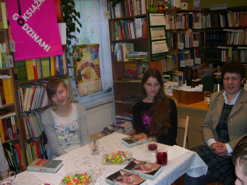 3 osoby siedzą przy stoliku, rozmawiają między sobą, na stole leżą omawiane książki, stoją szklanki z napojami i talerzyki z ciastkami.