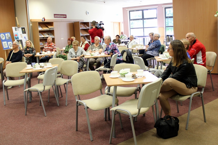 Publicznosć zebrana na spotkaniu, widać kobiety i mężczyzn w różnym wieku, siedzących przy stolikach