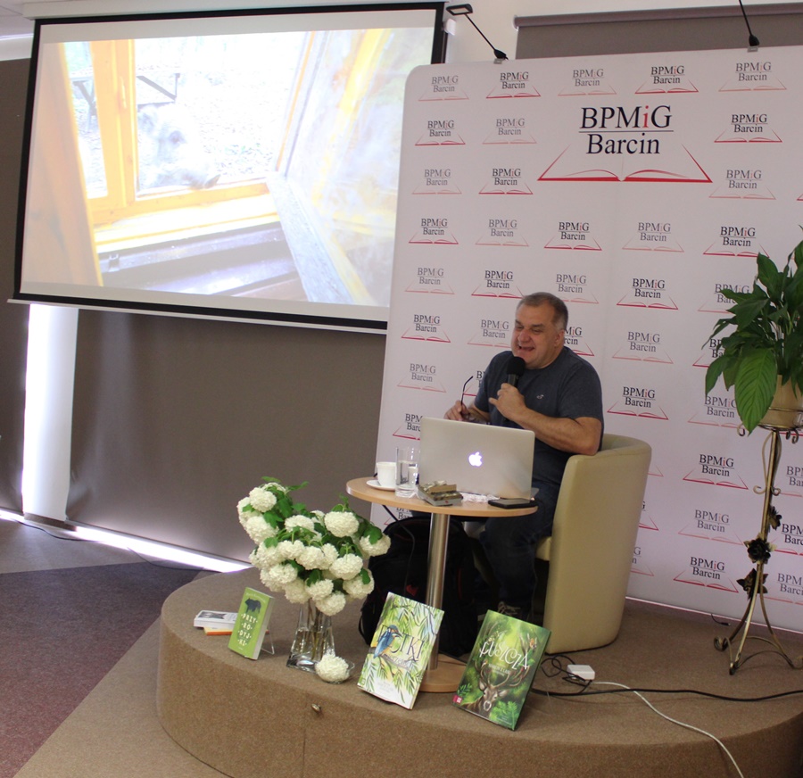 Pan Marcin siedzi w fotelu na scenie, przed nim stoi stolik z laptopem, na podeście ustawione są również książki jego autorstwa, na ekranie projektora wyświetlane jest zdjęcie z dzikiem zaglądającym przez okno.