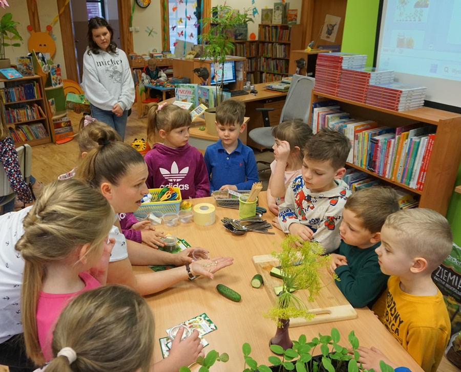 dzieci przyglądają się nasionkom które na dłoni trzyma bibliotekarka prowadząca zajęcia