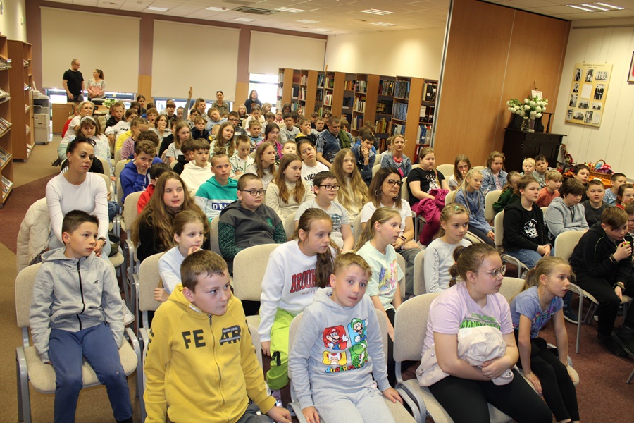 Kilkadziesiąt dzieci zebranych na spotkaniu razem z nauczycielami siedzą na krzesłach, patrzą przed siebie lub w stronę aparatu.
