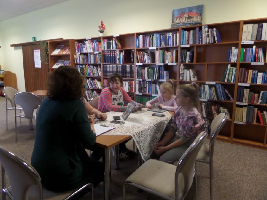 bibliotekarka i czwórka dzieci siedzą na krzesłach ustawionych dookoła stołów i rozmawiają