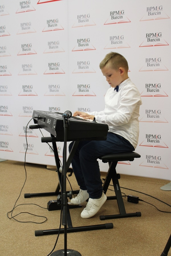 Młody chłopczyk ubrany na galowo siedzi na stołku i gra na instrumencie klawiszowym