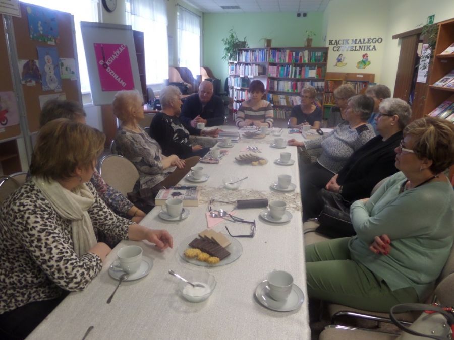 12 osób siedzących przy stołach w czasie spotkania rozmawia między sobą, na stołach widać poczęstunek w formie ciastek, kawy i herbaty.