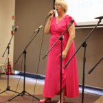 Nell Polgert-Martwijczuk (wysoka blondynka z krótkimi włosami ubrana w różową suknię) w czasie występu stoi przy mikrofonie i śpiewa przed cebraną publicznością