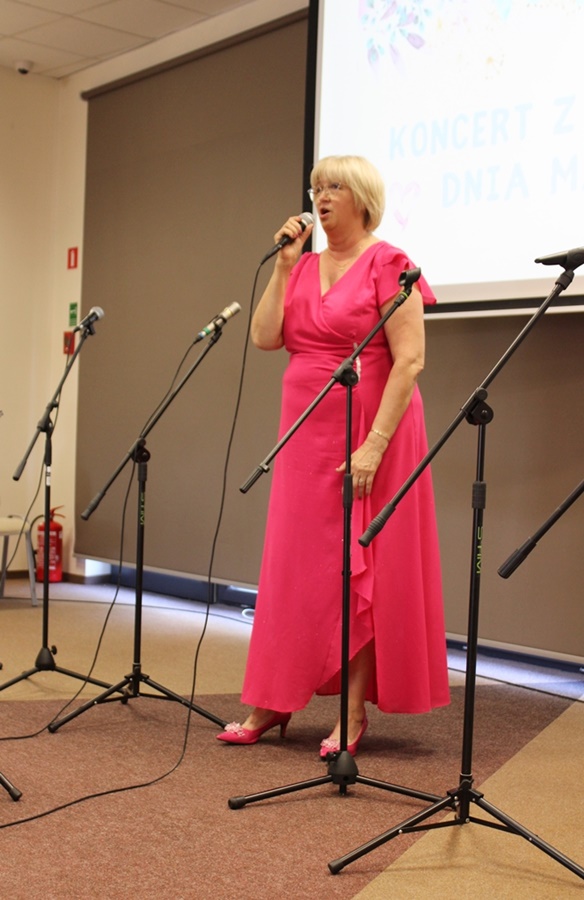 Nell Polgert-Martwijczuk (wysoka blondynka z krótkimi włosami ubrana w różową suknię) w czasie występu stoi przy mikrofonie i śpiewa przed cebraną publicznością