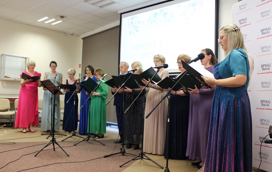 Zdjęcie występującego chóru, 12 osób ubranych na galowo stoi przy mikrofonach w rękach trzymają teczki z tekstami utworów które śpiewają.