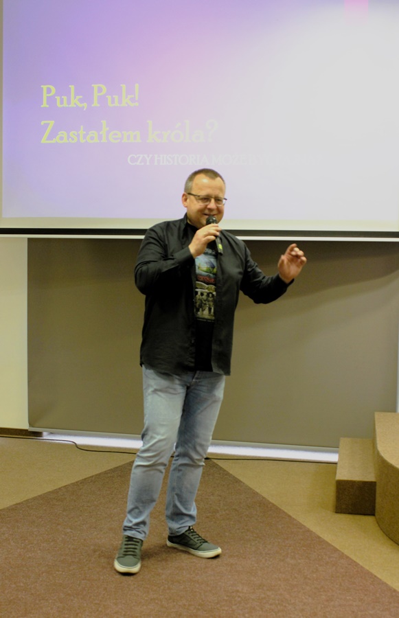 Uśmiechnięty Marcin Przewoźniak stoi z mikrofonem w ręce, za nim wyświetlana jest prezentacja multimedialna
