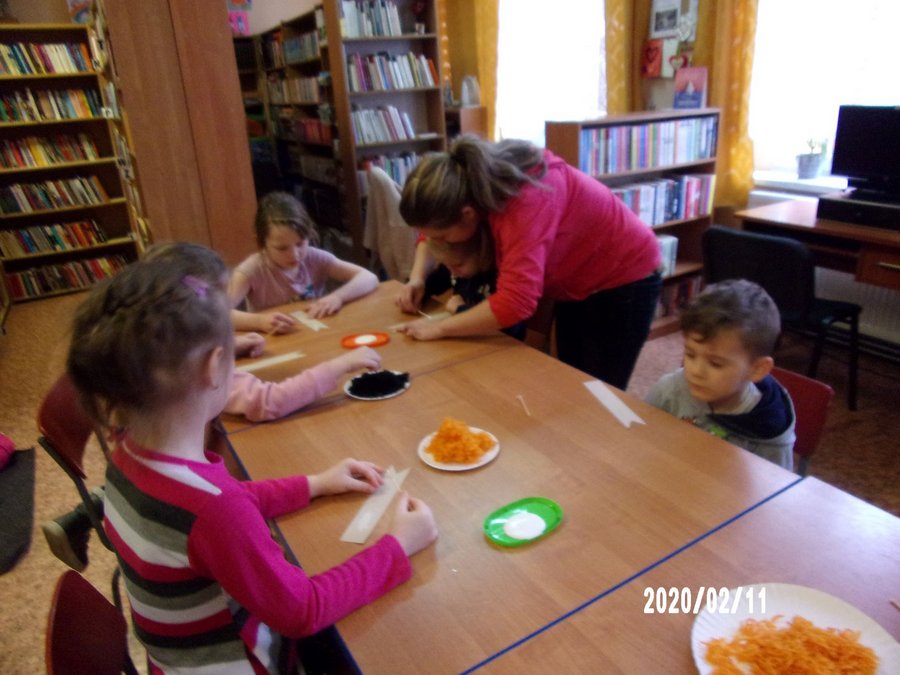 5 dzieci siedzi przy stołach, wolontariuszka pomaga w czymś jednemu z dzieci, na talerzykach leżą czarne i pomarańczowe nitki potrzebne do zajęć, przed dziećmi leżą tekturowe zakładki w kształcie głowy kota.