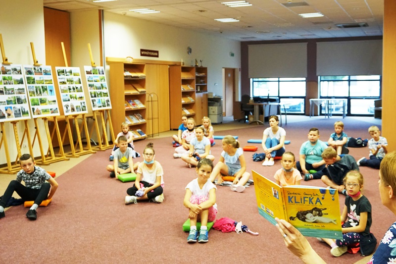 Zebrane dzieci siedzą na kolorowych poduszkach i słuchają pani bibliotekarki.