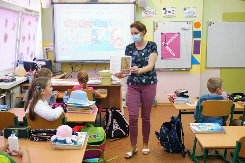 Prowadząca spotkanie bibliotekarka przechadza się po sali, pokazując dzieciom zawartość książki.