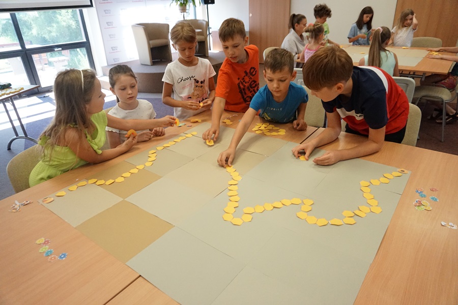 Grupa dzieci układających na szarych tekturach ścieżkę z żółtych kawałków papieru w kształcie plastra miodu