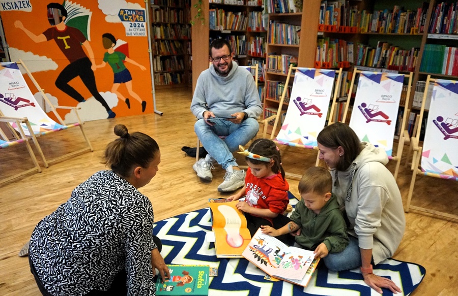 2 dzieci razem z matką siedzą na kocu na podłodze i oglądają książki, tata dzieci siedzi na leżaku i trzyma książkę którą bedzie czytał, z boku kuca bibliotekarka