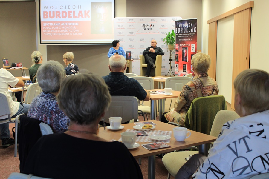 Dyrektorka biblioteki razem z zaproszonym gościem siedzą w fotelach na scenie przed zebraną publicznościa i rozmawiają o twórczości autora