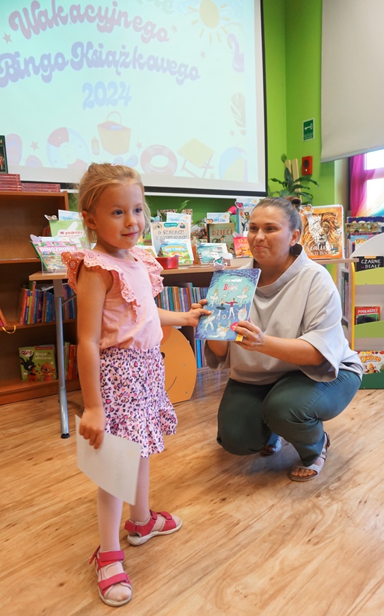 Dziewczynka ubrana na różowo odbiera z rąk bibliotekarki dyplom i książkę