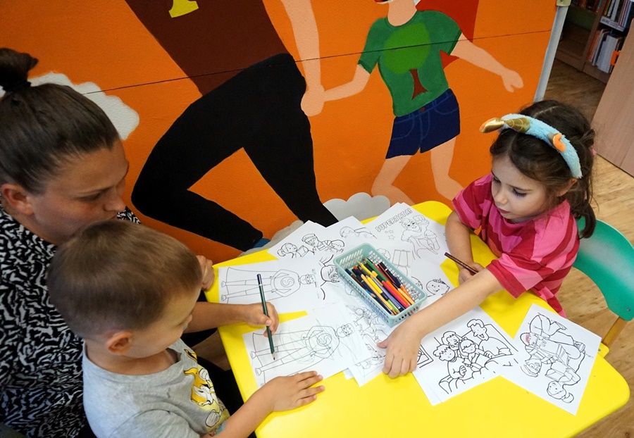 2 dzieci siedzi przy stoliku i koloruje kolorowanki, obok nich kuca bibliotekarka