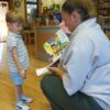 Chłopiec ubrany na biało-niebiesko odbiera nagrodę książkową z rąk bibliotekarki, która przy nim kuca.