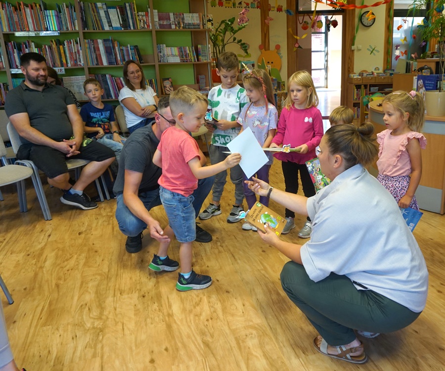 chłopiec w różowej koszulce i dżinsowych spodenkach odbiera dyplom i nagrodę od bibliotekarki, obok chłopca kuca jego ojciec