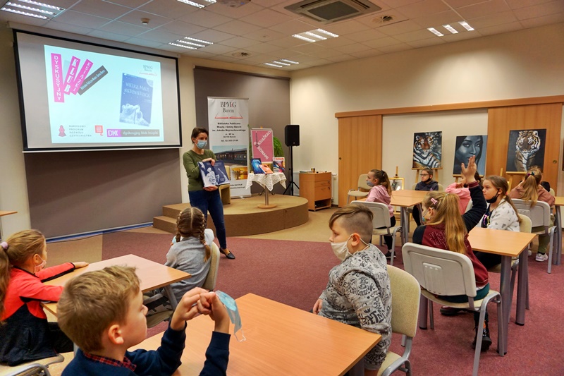 Bibliotekarka prowadząca spotkanie pokazuje dzieciom siedzącym przy stolikach otwartą książkę.