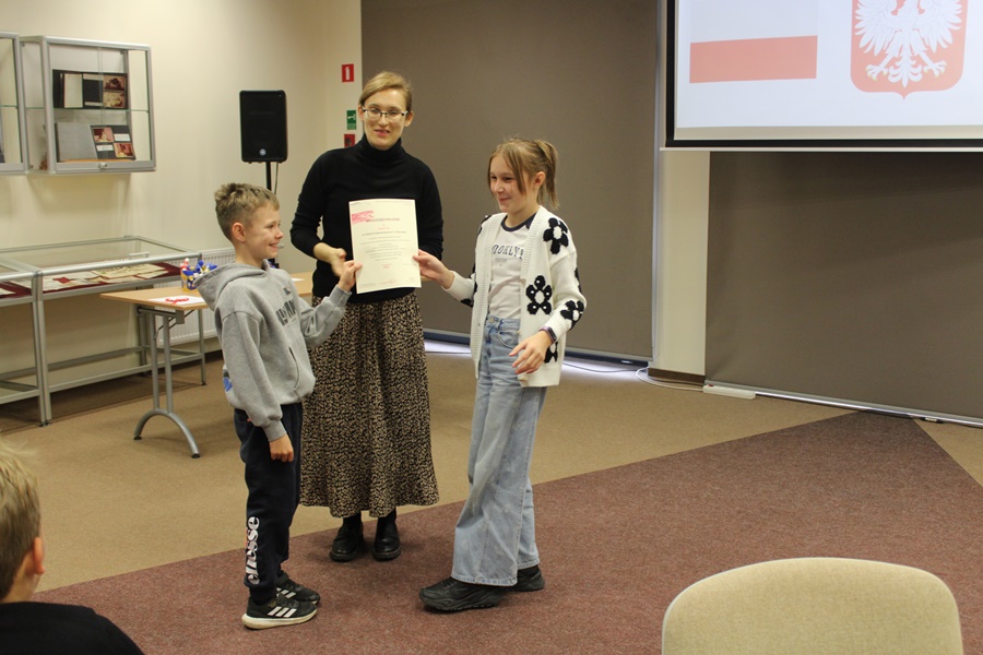 Bibliotekarka prowadząca zajęcia wręcza dyplom za udział na ręce dwójki dzieci.