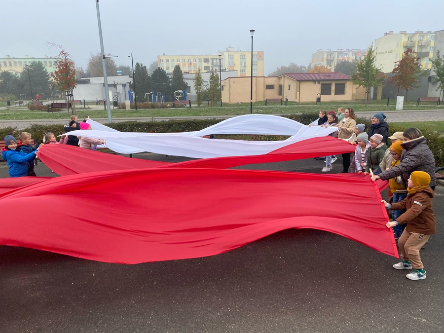 Dzieci i opiekunowie pzred budynkiem biblioteki rozciągają pasy białego i czerwonego materiału