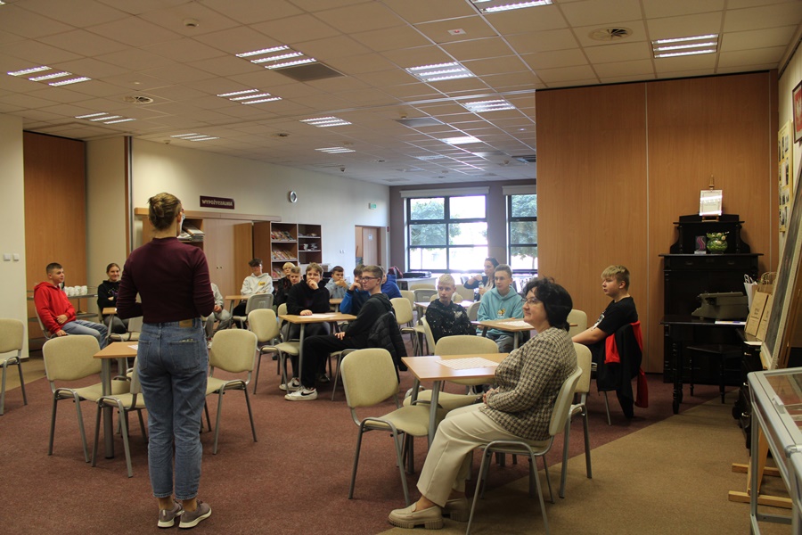 Wszyscy uczestnicy razem z dyrektorką biblioteki i koordynatorką projektu zebrani w czytelni biblioteki, jedna osoba stoi przed grupą i tłumaczy zasady zorganizowanego wydazrenia