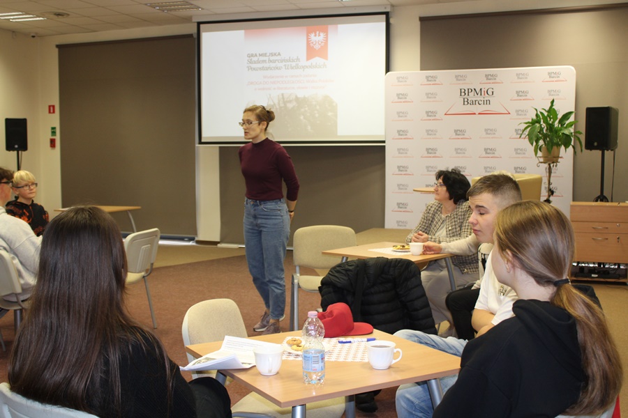 zdjęcie z miejsca siedzenia uczestników, w kadrze na pierwszym planie widać kilkoro z uczestników, głębiej stoi koordynatorka projektu i tłumaczy zasady zabawy, w tle widoczny jest plakat promujący dydarzenie wyświetlany na ekranie projektora