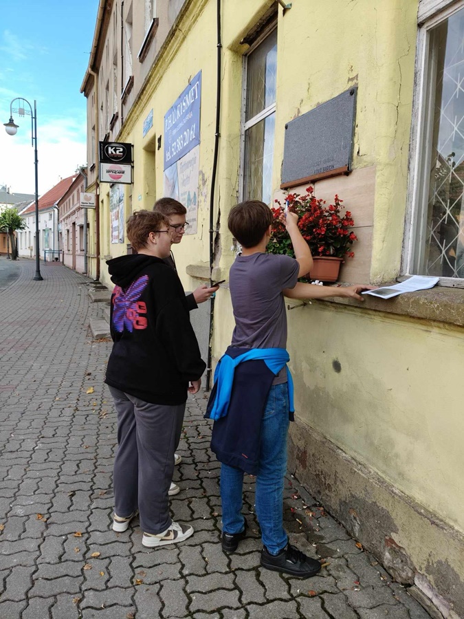 3 osoby stojące przed budynkiem na którym wisi tablica pamiątkowa, osoby rozmawiają między sobą, jedna zapisuje coś na kartce papieru